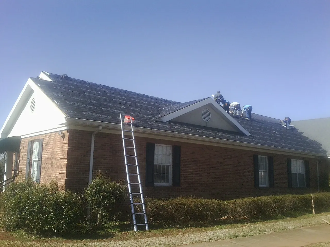 Crew tearing shingles off a brick ranch roof with a ladder set — Emergency Roof Repair work by our crew in Ogema