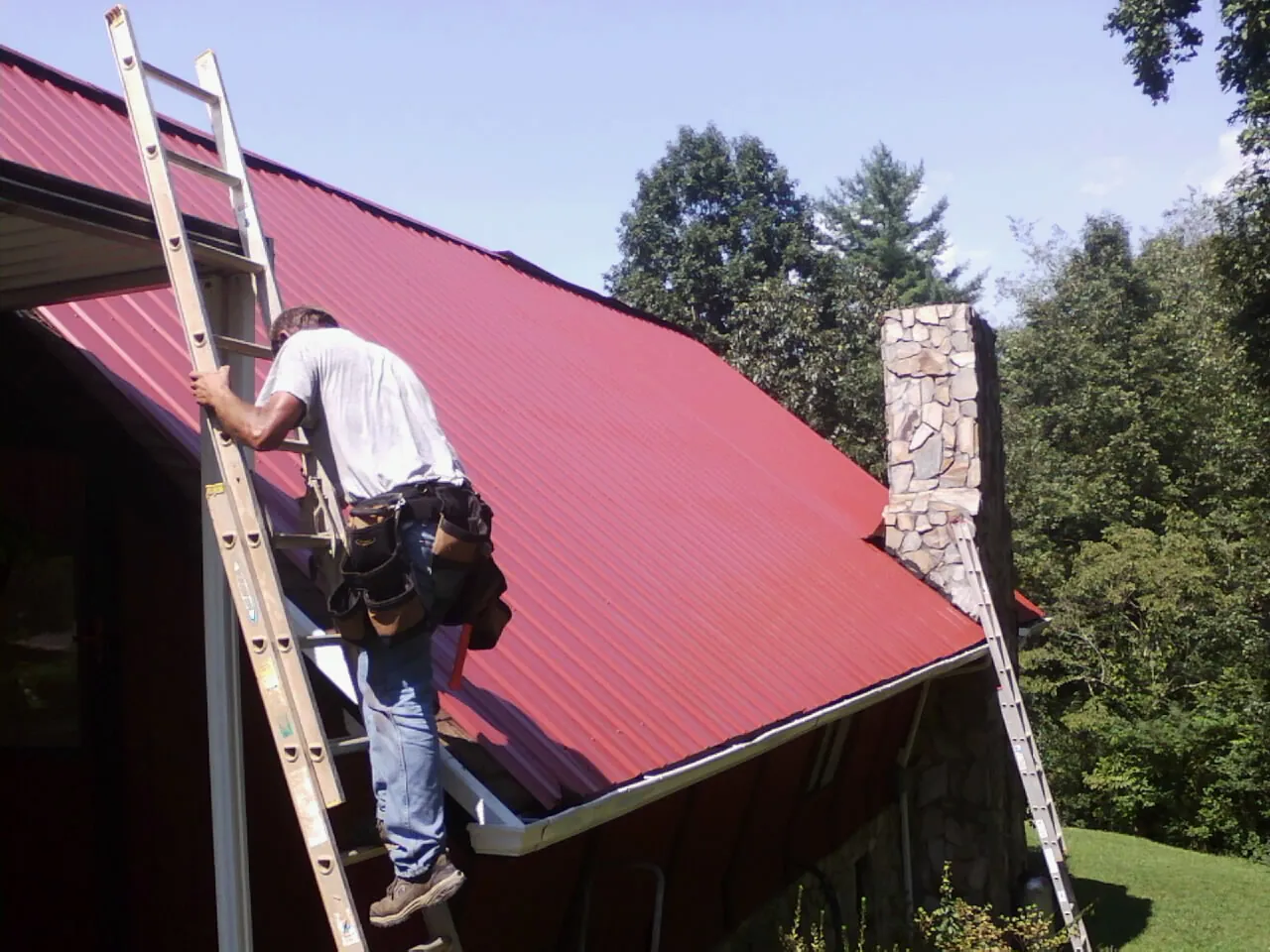 Roofer on a ladder beside a red metal roof and stone chimney — Soffit & Fascia Repair by Roofing Brothers in Ogema